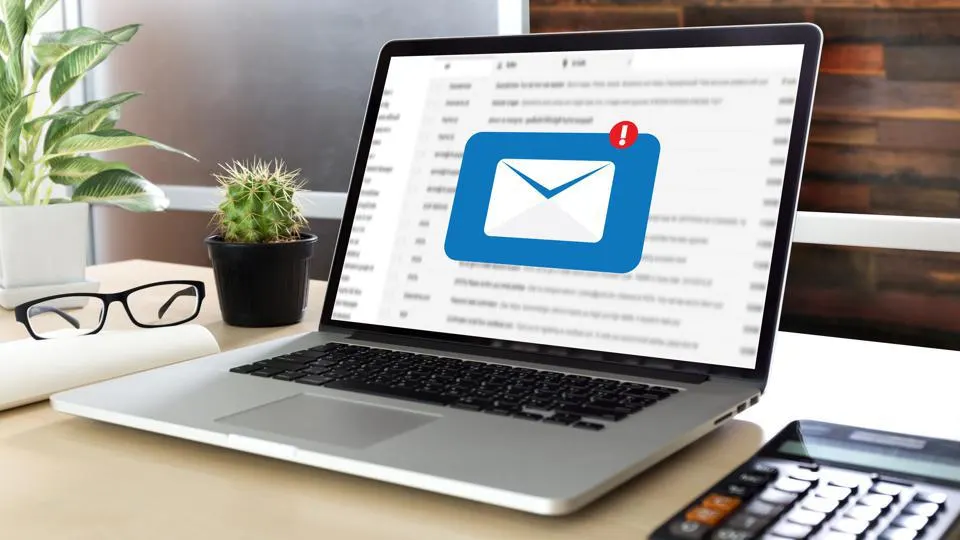 logo d'un Email bleu s'affichant sur l'écran d'un ordinateur portable posé sur un bureau élégant avec sur la gauche des plantes et une paire de lunettes et sur la droite une calculatrice